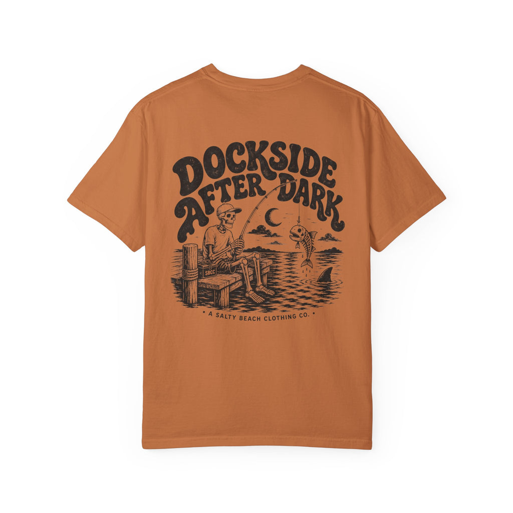 An orange short-sleeve t-shirt with a vintage-style black graphic on the front. The graphic features the text 'DOCKSIDE AFTER DARK' in a stylized font above an illustration of a man fishing from a dock at night, with a fish jumping out of the water under a crescent moon.