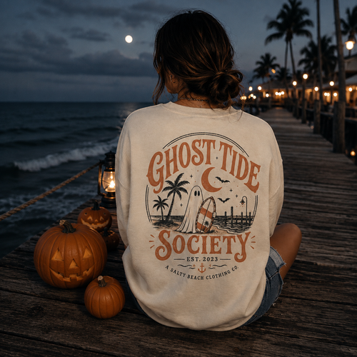 A woman sits on a wooden boardwalk at dusk facing the ocean, wearing a cream-colored graphic sweatshirt with a 'Ghost Tide Society' Halloween design. Jack-o'-lanterns are on the deck beside her, and a pier stretches into the background with warm lights and palm trees.