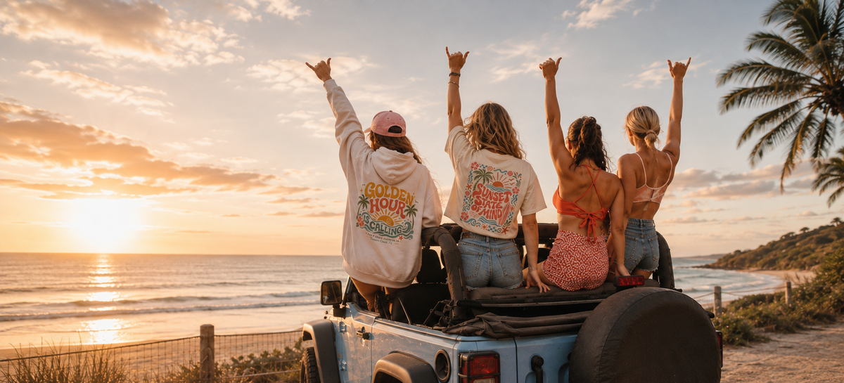 Four women sitting in the back of a light blue open-top vehicle on a beach at sunset, facing the ocean with raised hands. The vehicle has a large 'Cruel Hound' graphic on the back seat and a 'SALTY' license plate.