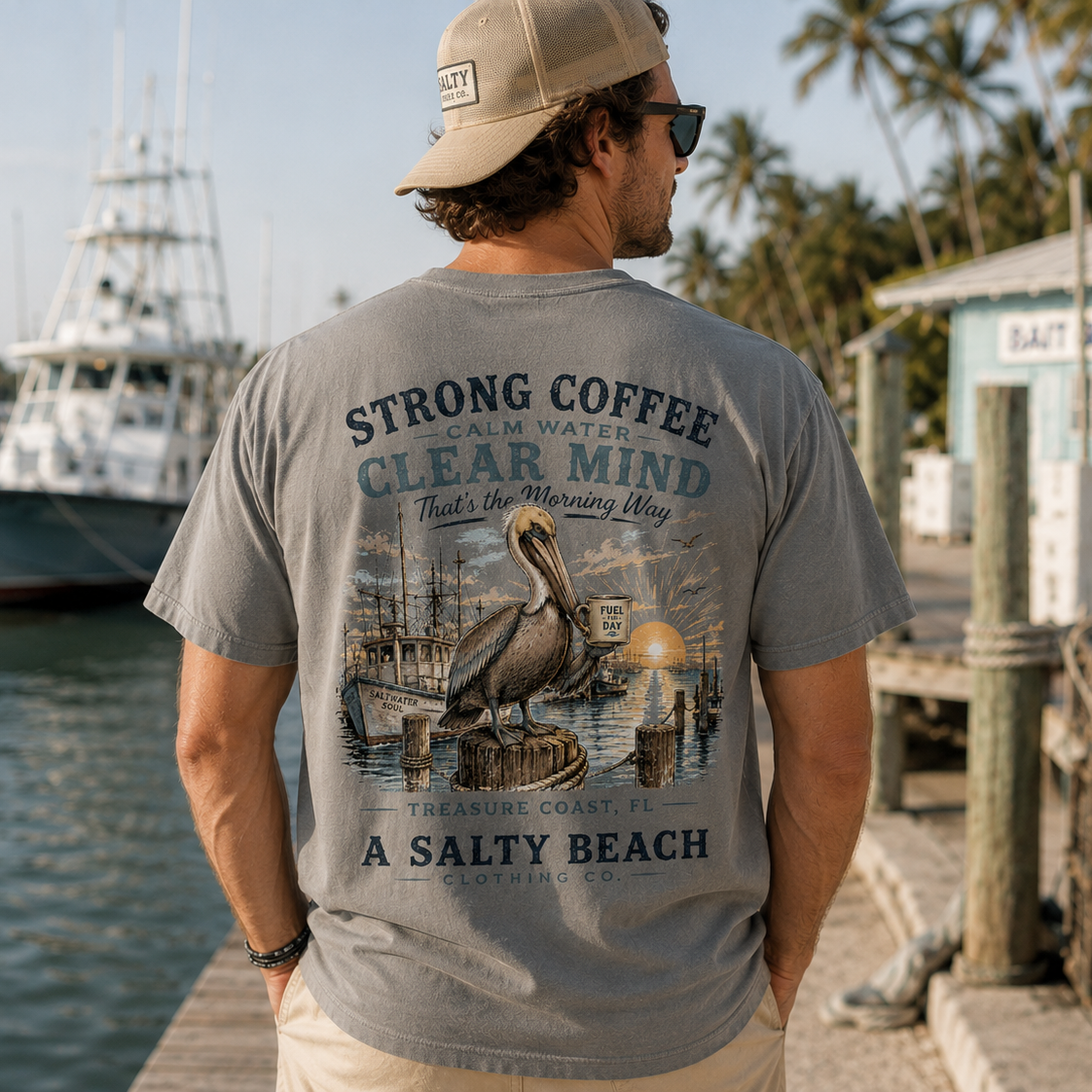 A man standing on a dock wearing a gray graphic t-shirt with a vintage-style nautical design featuring a pelican and the text 'STRONG COFFEE PALM WATERS CLEAR MIND That's the Morning Way Treasure Coast: FEEL A SALTY BEACH'.