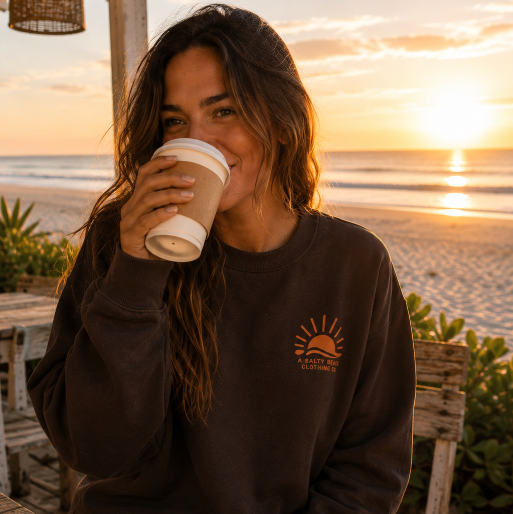 A woman sitting on a beach patio at sunset wearing a dark brown crewneck sweatshirt with a small orange sun graphic on the left chest, holding a white coffee cup.