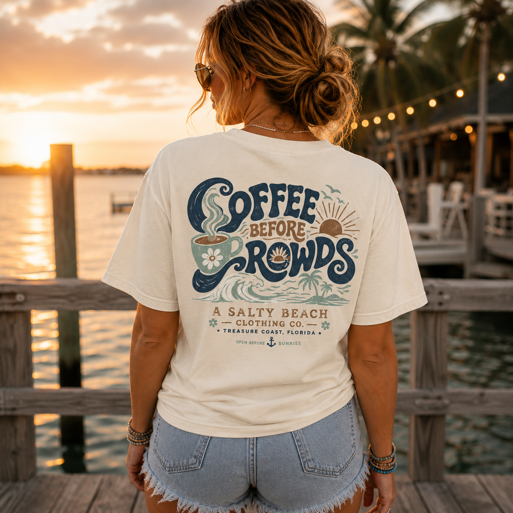 A woman stands on a wooden dock at sunset wearing a cream graphic T-shirt with a large back print reading 'Coffee Before Rows' and 'A Salty Beach Clothing Co.' in navy, tan, and teal tones, paired with light denim shorts.