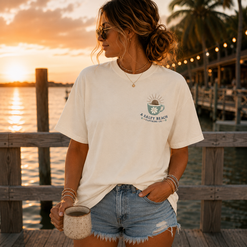 A woman stands on a wooden dock at sunset wearing an oversized off-white graphic T-shirt with a coffee cup design and the text 'A SALTY BEACH CAYMAN ISLAND'.