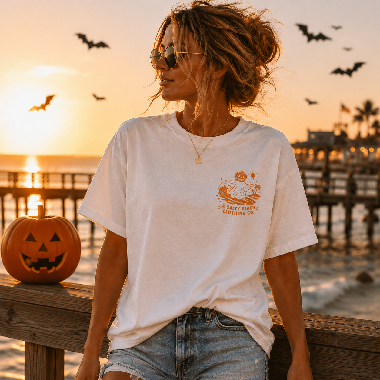 A woman wearing a white graphic t-shirt with a pumpkin and ghost design, set against a warm sunset beach background with a jack-o'-lantern on the railing.