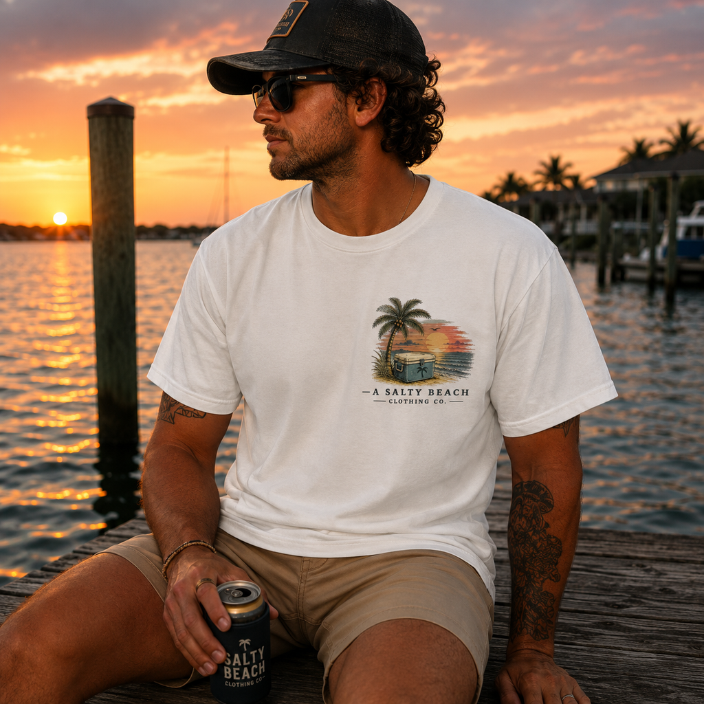A man sitting on a wooden dock at sunset wearing a white graphic t-shirt with a 'Salty Beach' palm tree and beach graphic, khaki shorts, and a black cap.