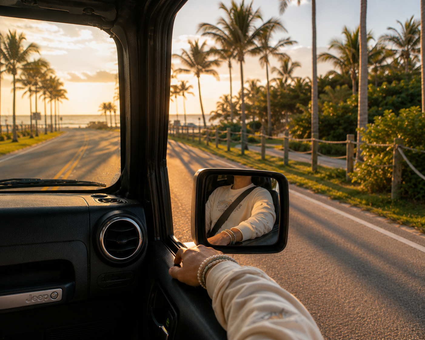 A person driving a vehicle on a tropical coastal road at sunset, with palm trees, ocean, and the driver's arm visible in the side mirror.
