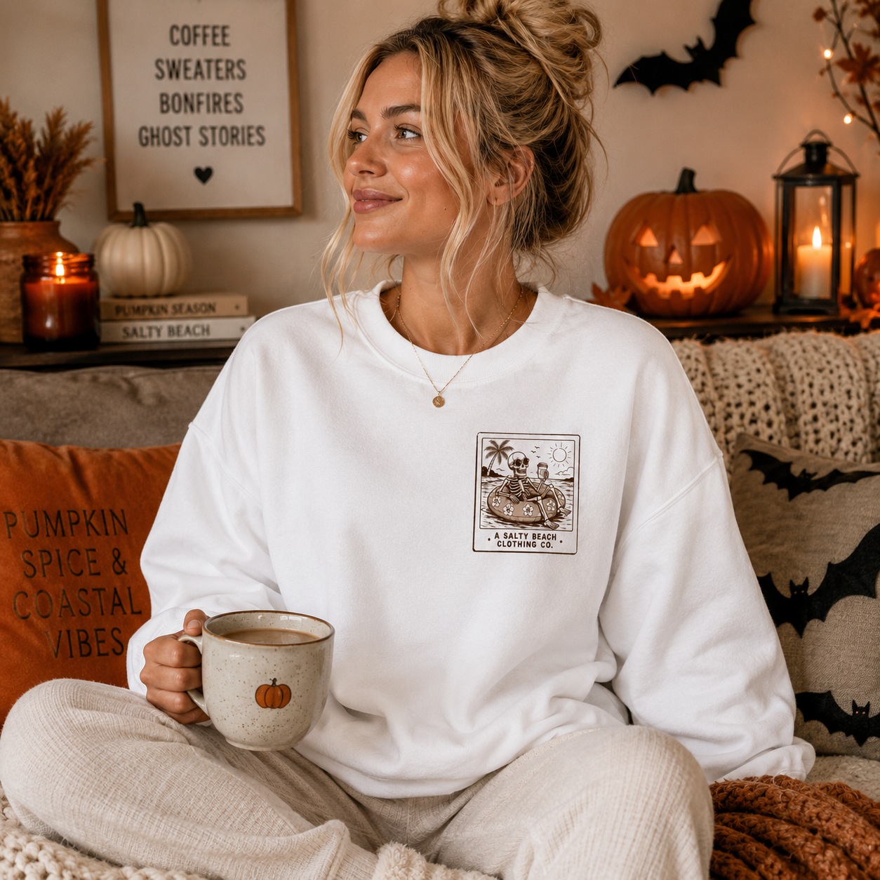 A woman sits cross-legged on a couch in a cozy living room, wearing a white crewneck sweatshirt with a brown vintage-style graphic on the left chest that reads 'A South Boston Affairine Co.' She holds a ceramic mug, with pumpkins, candles, and Halloween decorations in the background.