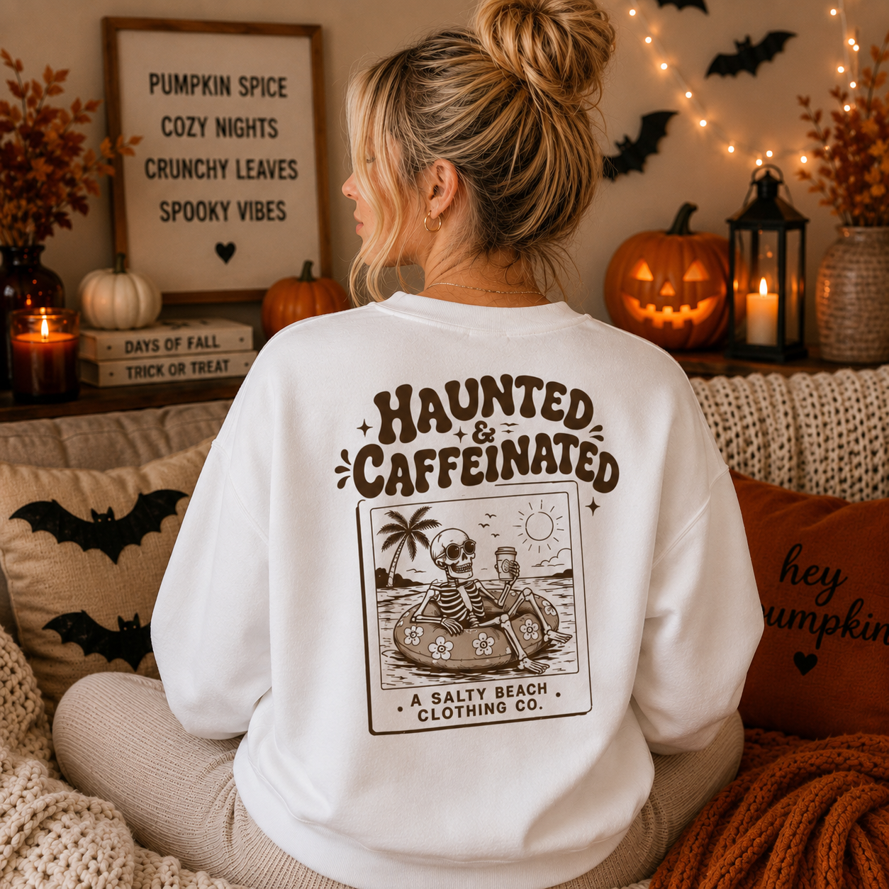 A woman sits on a couch wearing a white sweatshirt with a large brown graphic on the back that reads 'Haunted & Caffeinated' and shows a cartoon skeleton in a floatation ring. The scene is decorated for Halloween with pumpkins, string lights, and cozy pillows.