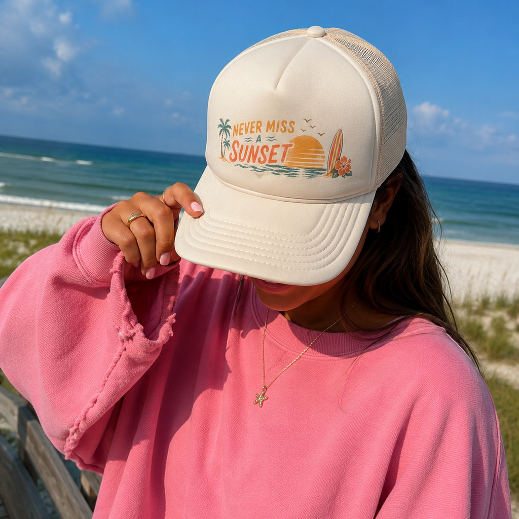 A person wearing a cream-colored trucker hat with a 'Never Miss the Sunset' graphic, paired with a pink sweatshirt, standing on a beach dune with the ocean and sky in the background.